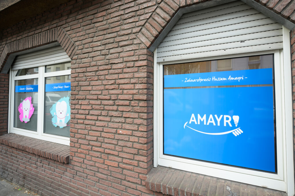 Fensterfront der Zahnarztpraxis Amayri in Duisburg mit Praxislogo und freundlichen Zahnillustrationen