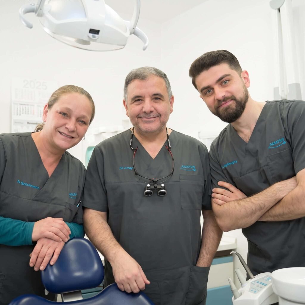 Drei Mitglieder des Teams der Zahnarztpraxis Amayri stehen lächelnd neben einem blauen Behandlungsstuhl.